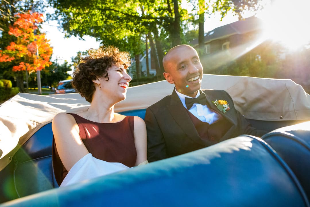 A bride and groom smile beneath the shining sun as they ride in the back of a convertible.