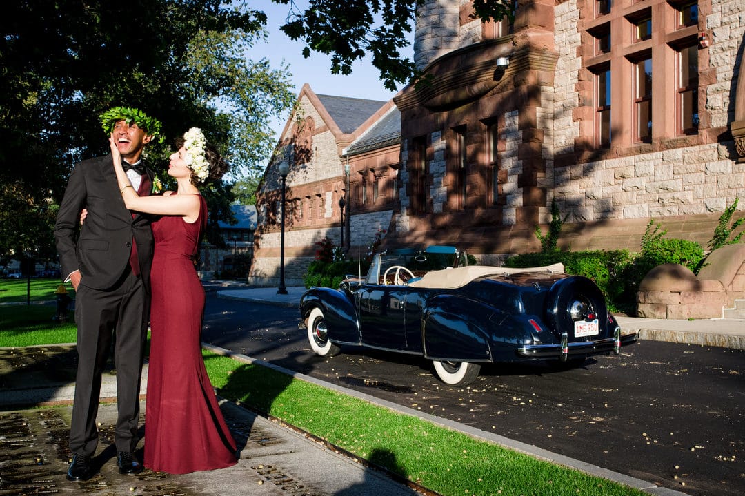 A bride wearing a maroon dress and her groom laugh as they embrace next to a blue antique Lincoln Continental convertible.