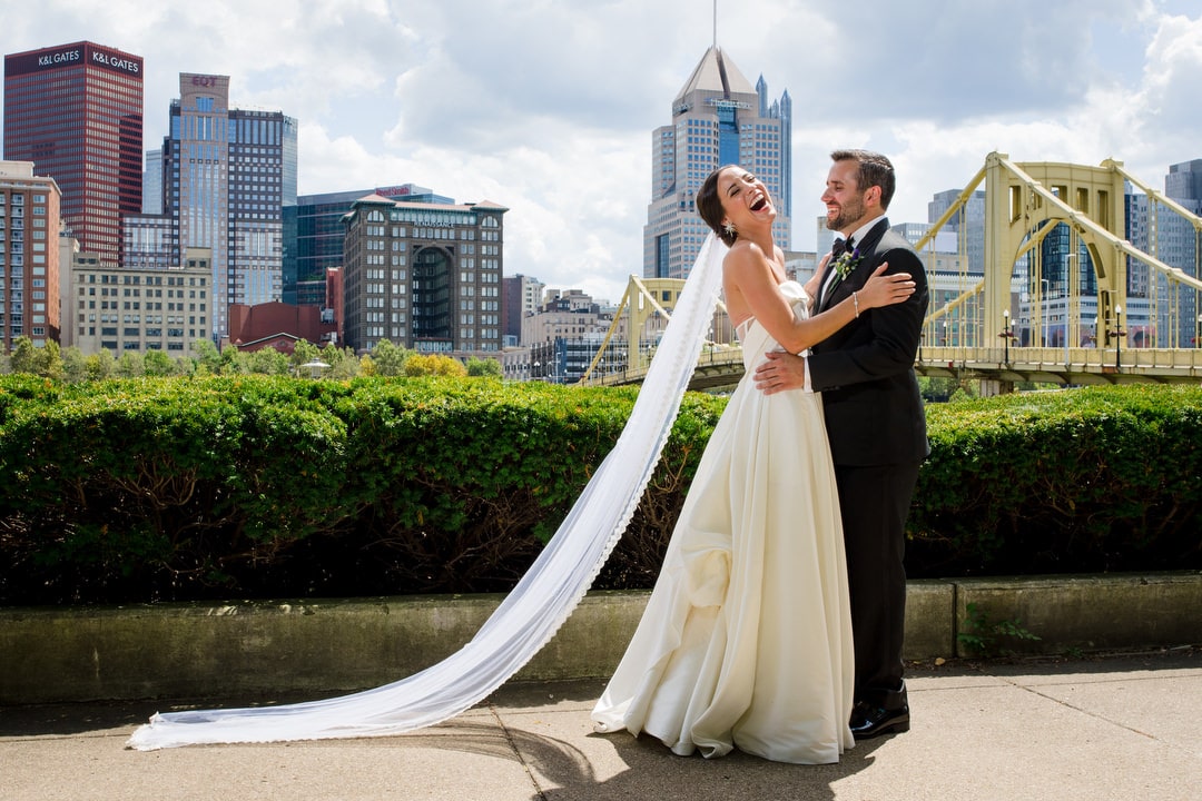 A bride wearing a long white veil holds her husband and laughs in front of the skyline of Pittsburgh and a yellow bridge.