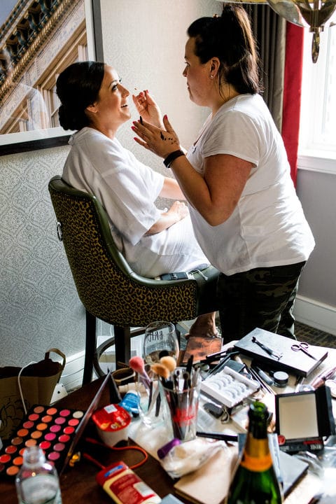 A bride sits on a tall chair as another woman applies makeup to her.