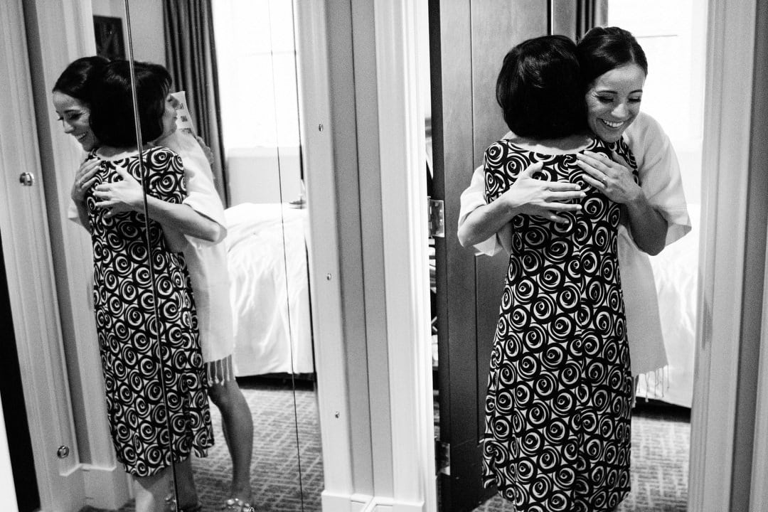 A bride to be in a bathrobe hugs her future mother in law. They're reflected in a mirror.