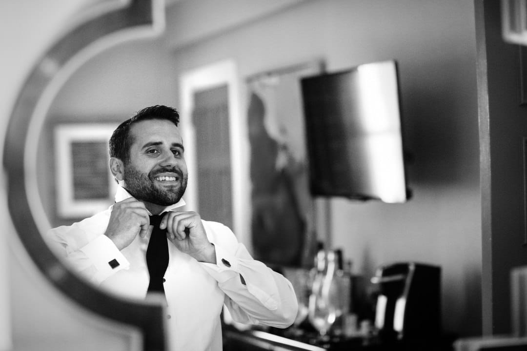 A groom with a dark beard ties his bow tie in a mirror.