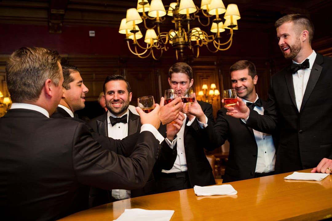 A group of men gathered around a wood bar hold glasses of whiskey up as they toast.