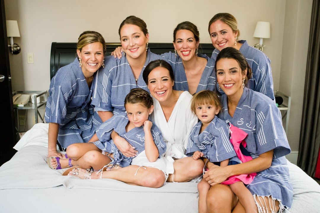 A group of bridesmaids wearing blue bath robes surround a bride wearing a white monogrammed robe.