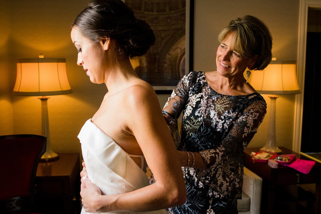 A bride holds her dress as her mother zips up the back in a hotel room.