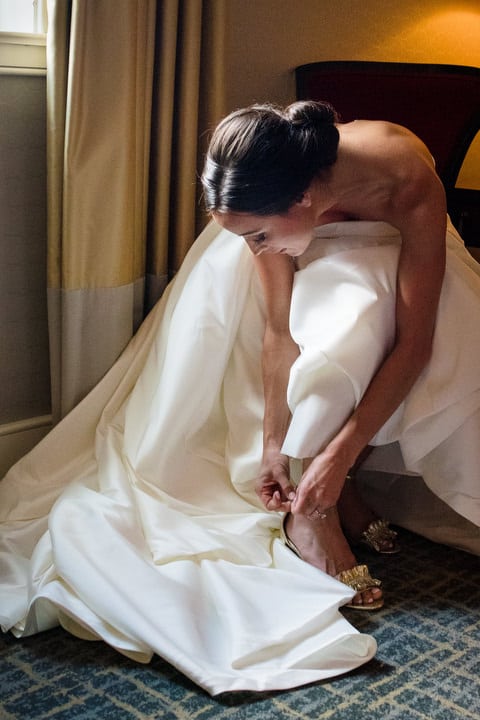 A bride bends over to buckle the strap on her shoe while sitting in a chair wearing a long white dress.