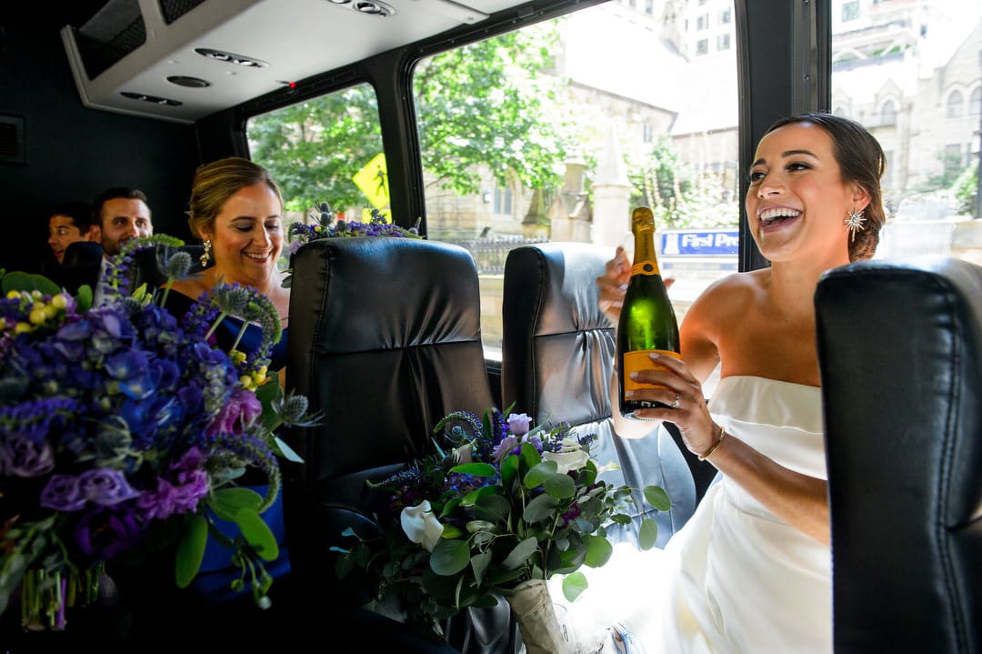 A bride smiles as she opens a bottle of champagne while riding on a bus with her bridal party.