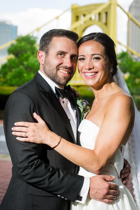 A bride and groom hold each other and smile as they stand in front of a yellow bridge in Pittsburgh.