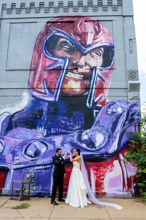 A bride and groom toast each other as they stand in front of a large mural of Magneto.