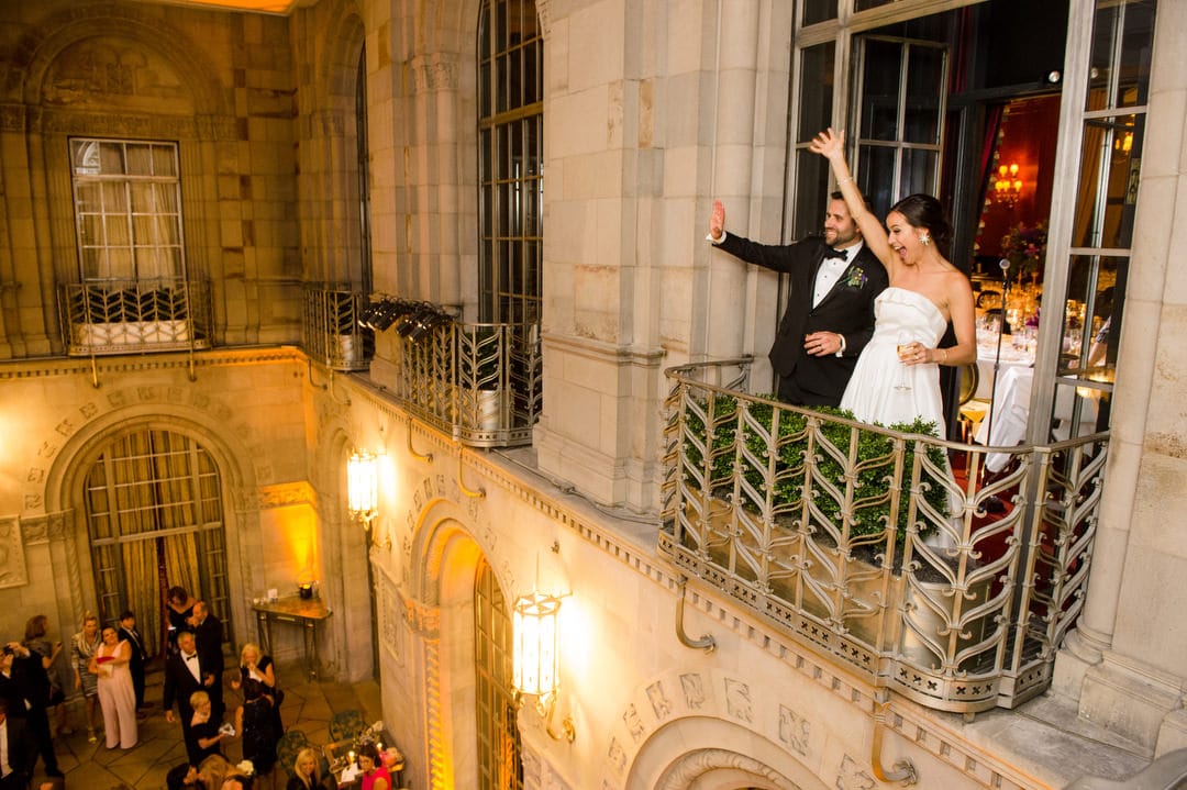 A bride and groom stand on a balcony and wave to their guests during their wedding at the Duquesne Club.