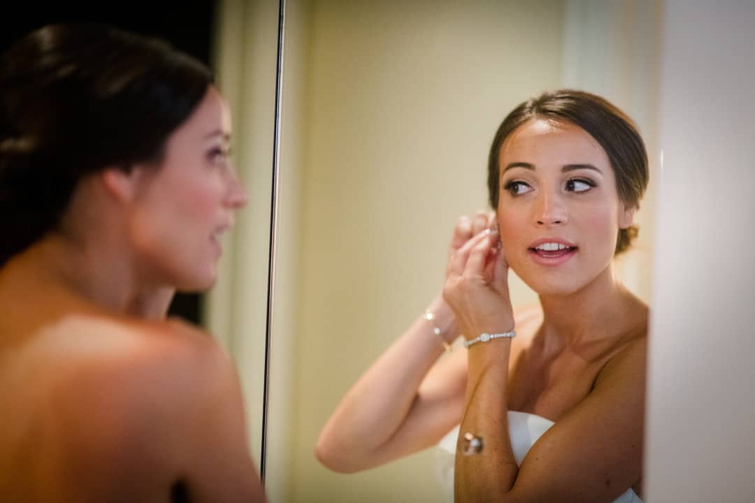 A bride looks into a mirror as she puts on one of her earrings.