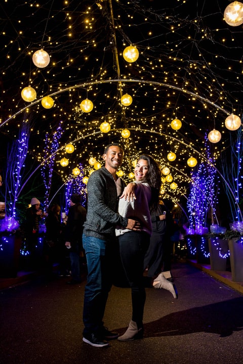 A young man and woman stand together beneath a tunnel of Christmas lights while she shows off her engagement ring.