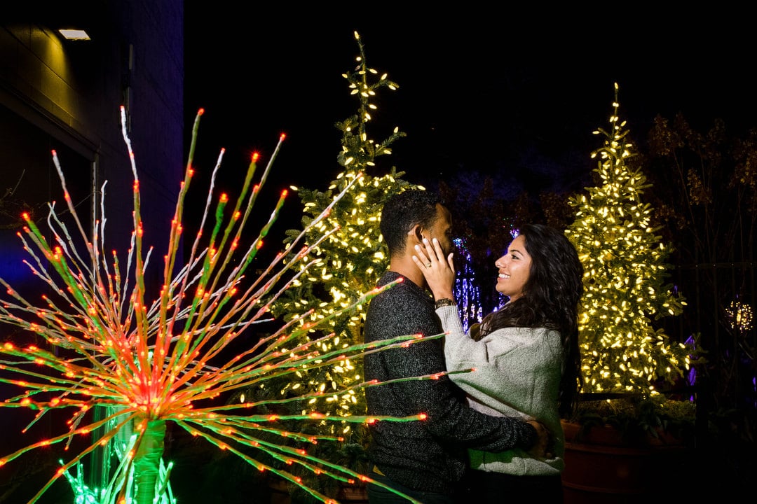 A black haired young woman brushes the face of her fiance as they stand together amongst Christmas light decorations.