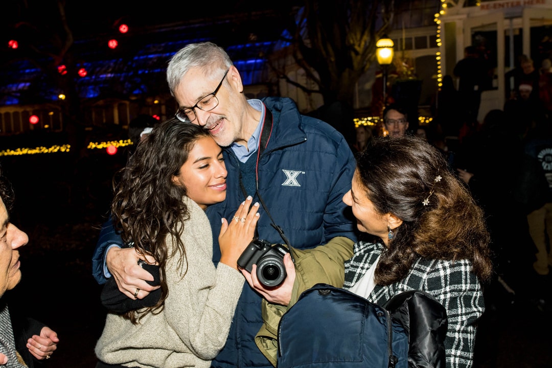A young woman in a gray sweater hugs her father as her mother looks on.