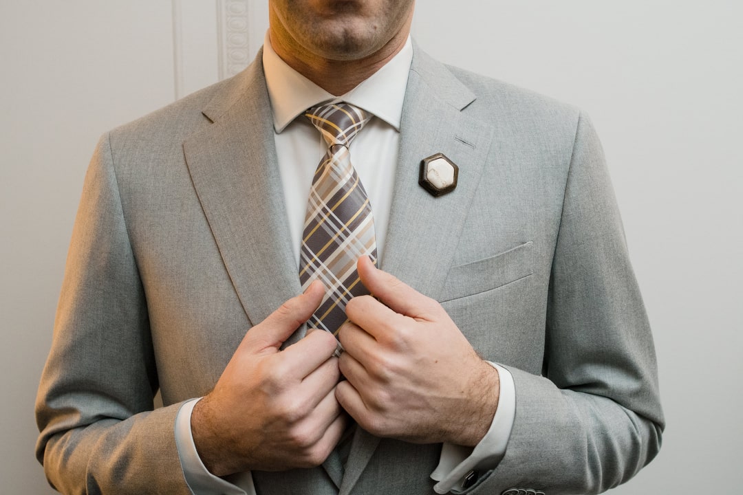 A man holds the lapels of his gray suit. He's wearing a brown patterned tie and a hexagon pin.