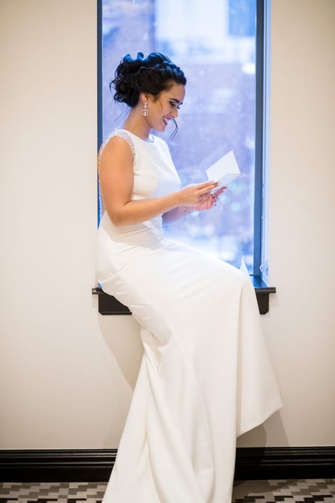 A bride in a white dress smiles as she sits in a window and reads a card at the Hotel Monaco Pittsburgh.