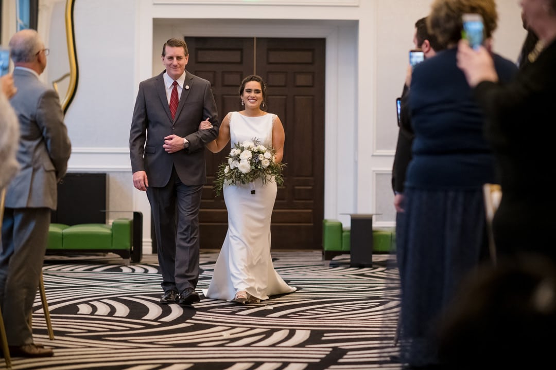 A smiling bride walks with her father into her winter wedding at the Hotel Monaco in Pittsburgh.