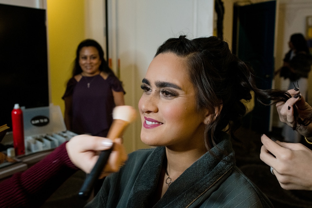 A woman smiles as a makeup brush applies blush to her face. An older woman looks on and smiles in the background.