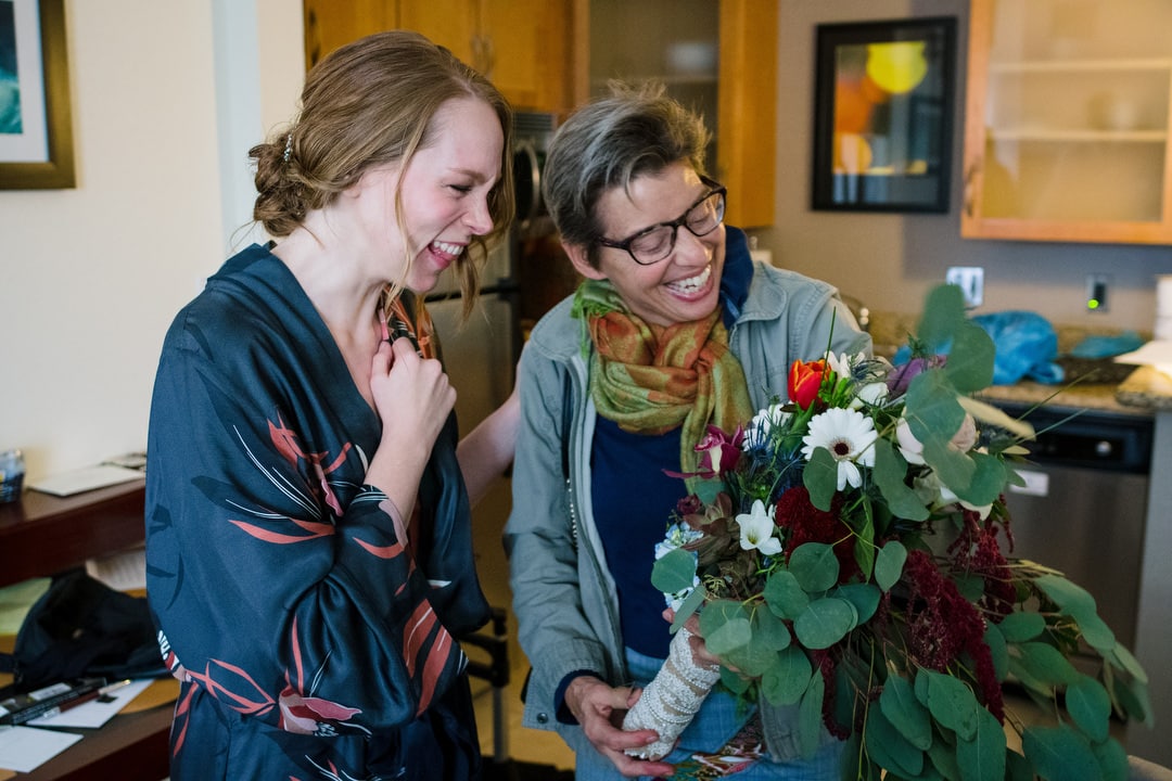 A woman wearing a dressing gown smiles at a bouquet held by another woman wearing glasses.
