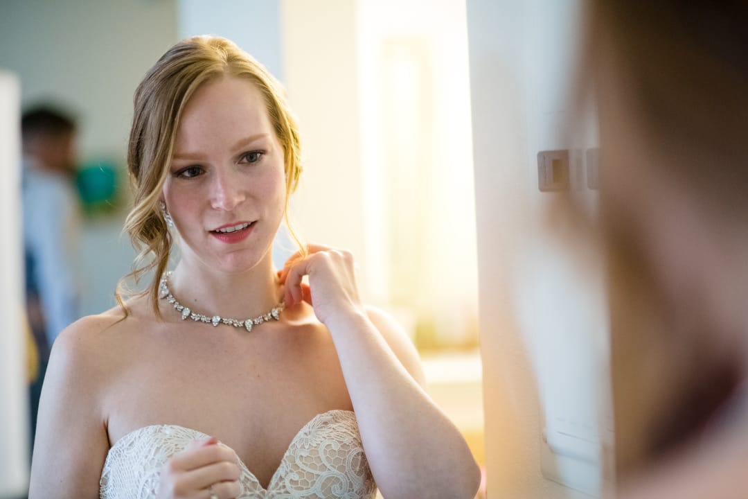 A bride wearing a white dress looks into a mirror as she adjusts her necklace.