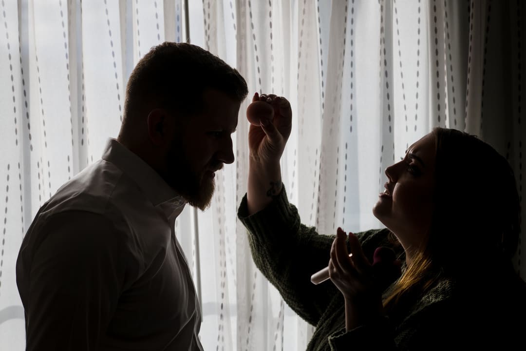 A silhouette of a makeup artist applying powder to the forehead of a bearded man.
