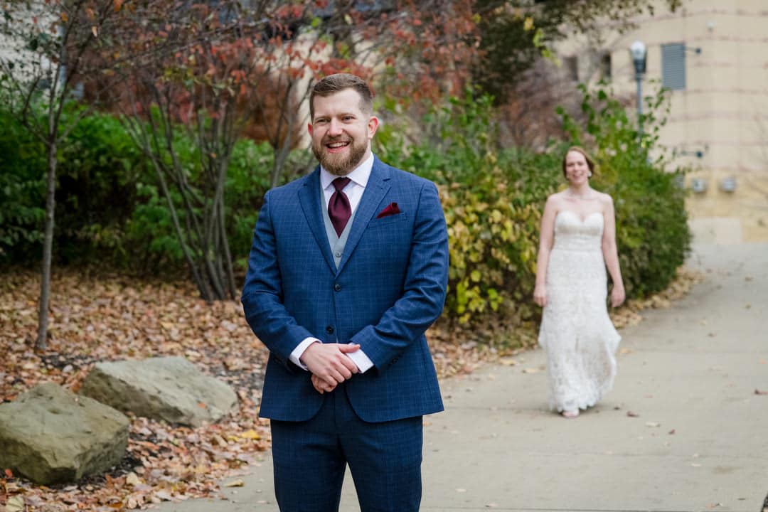 A groom wearing a blue suit smiles as his bride approaches him from behind on the path near PNC Park in Pittsburgh.