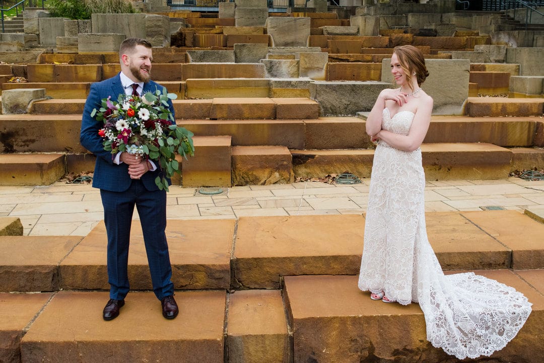 A groom holds his bride's bouquet as they stand on the water steps on the north shore of Pittsburgh.