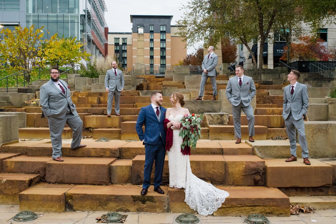 A bride and groom stand together on the water steps on the North Shore of Pittsburgh as their all-male bridal party in gray suits stands around them.