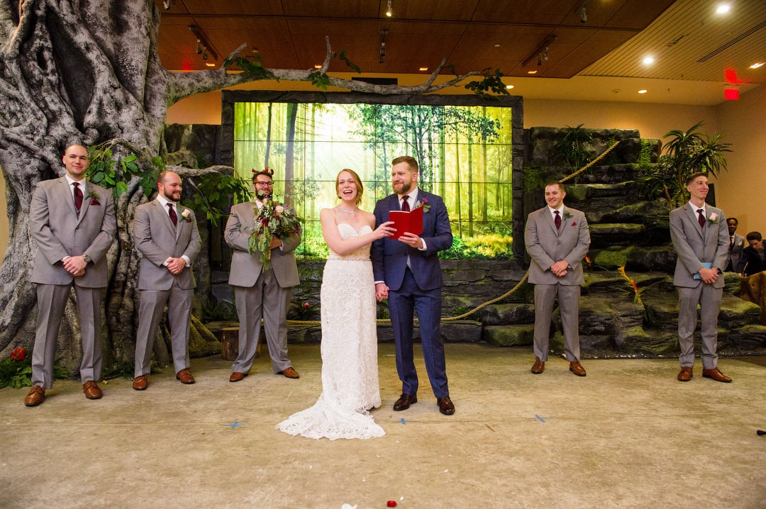A bride and groom stand together holding a red book as 5 men in gray suits stand around them during their Pittsburgh Aviary wedding.