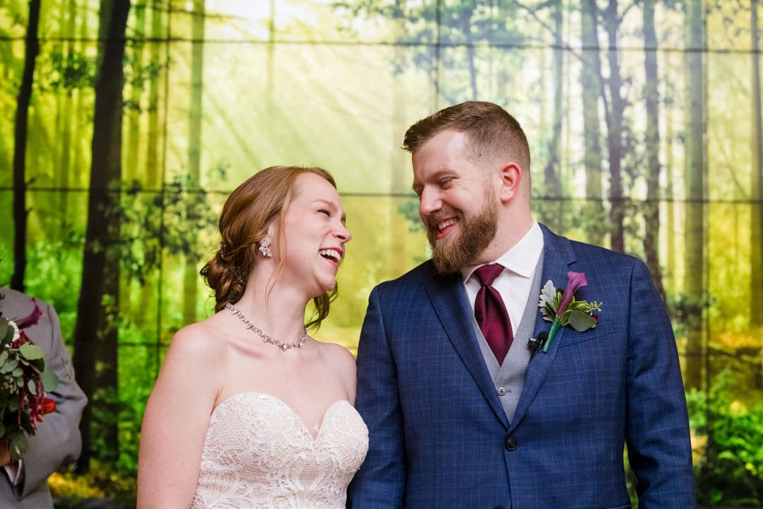 A bride and groom smile at each other as they stand in front of a forest image projected on a screen behind them.