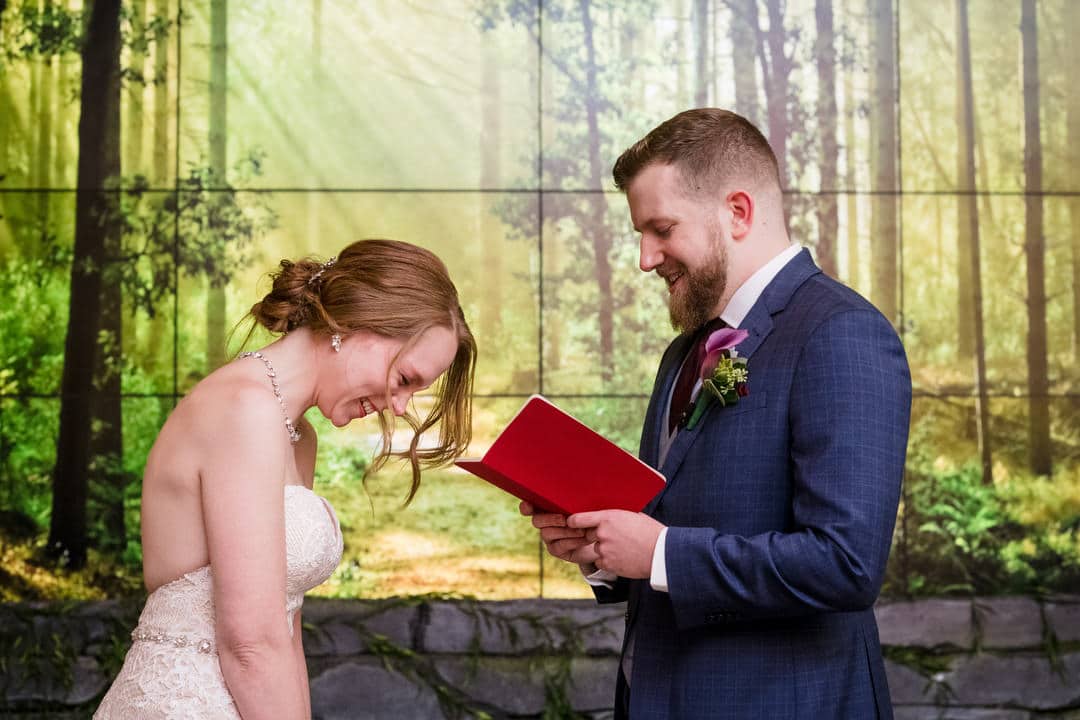 A bride laughs as her groom reads his vows out of a red booklet during their Pittsburgh Aviary wedding.