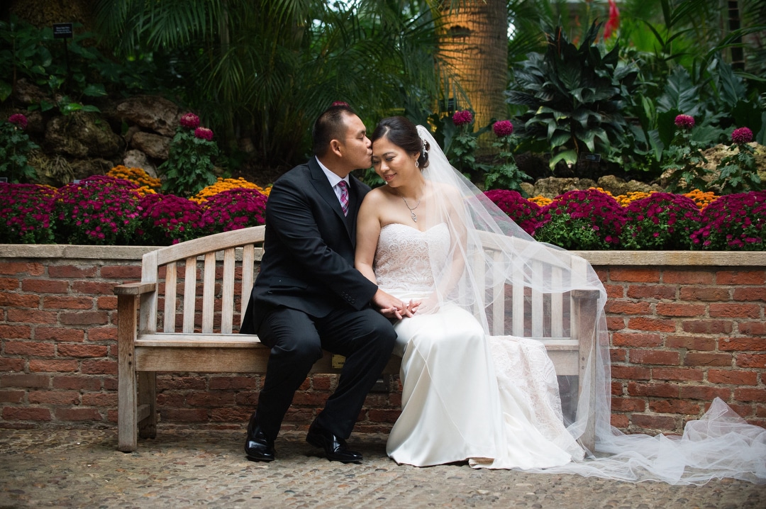 A groom kisses his bride's forehead as they sit on a bench in the Palm Court after their small wedding at Phipps.