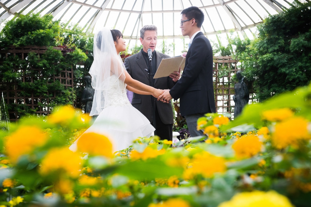 A bride and groom hold hands with yellow flowers in the foreground as a reverend performs a wedding ceremony in the broderie room during their small wedding at Phipps.