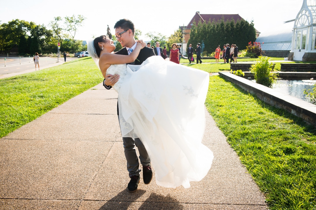 A groom carries his bride in his arms in front of Phipps Conservatory and Botanical Gardens.