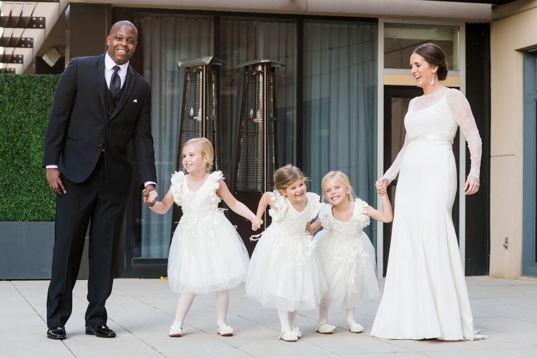 A bride and groom hold hands with their three flower girls while they all smile.