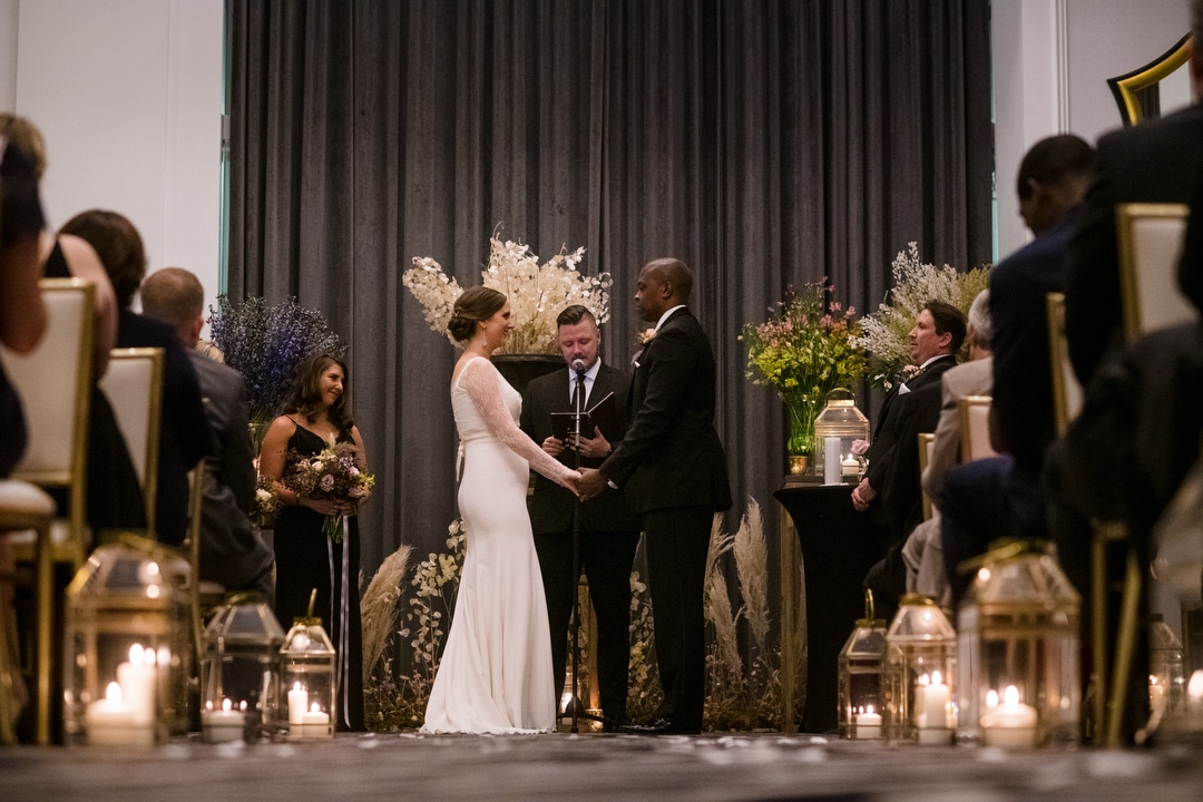 A bride and groom stand in front of gray curtains and large flower arrangements during their Hotel Monaco Pittsburgh wedding.