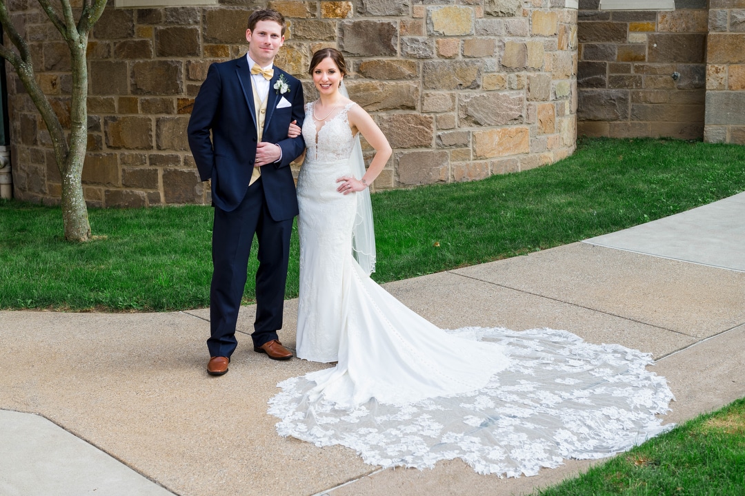 A bride and groom stand on a sidewalk with a stone wall behind them. Her dress is spread out next to her.