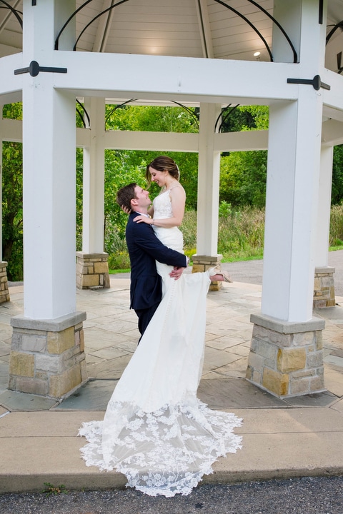 A groom lifts his bride up as her dress flows out around them beneath a gazebo.