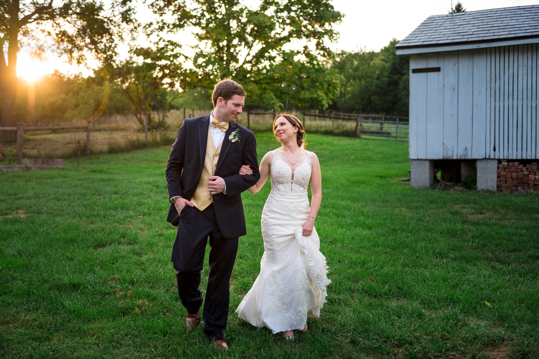 A bride and groom walk through a barn yard arm in arm as the sun sets behind them.
