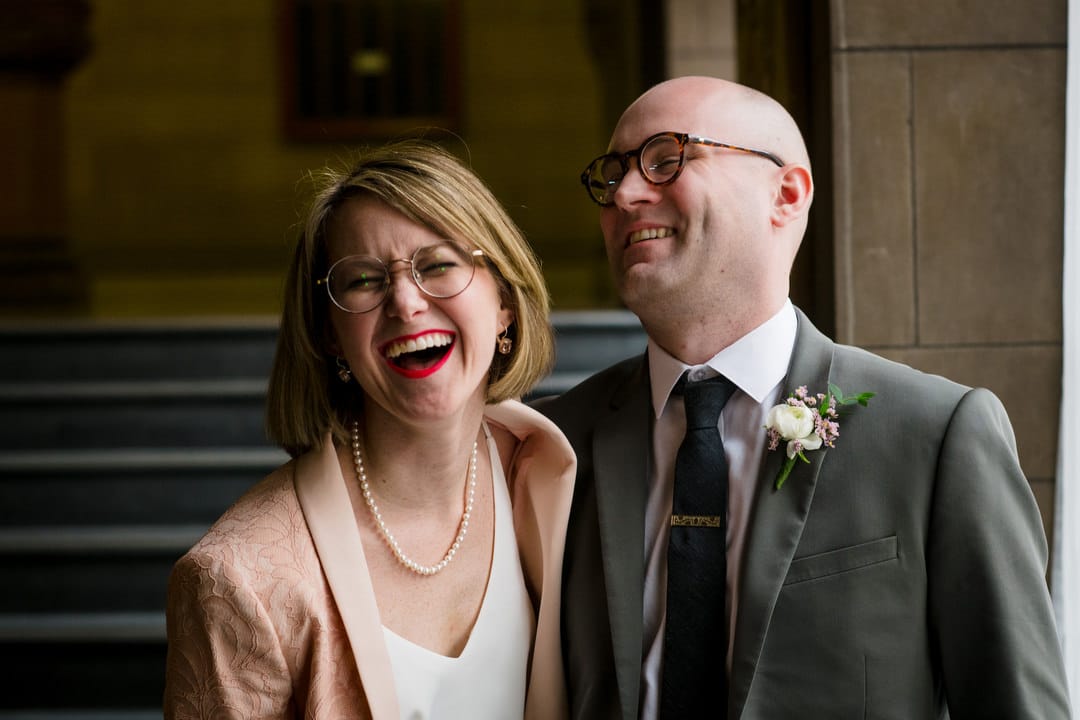 A bride wearing a pink pantsuit and a groom in a gray suit laugh on the stairs of the Allegheny County Courthouse in Pittsburgh.