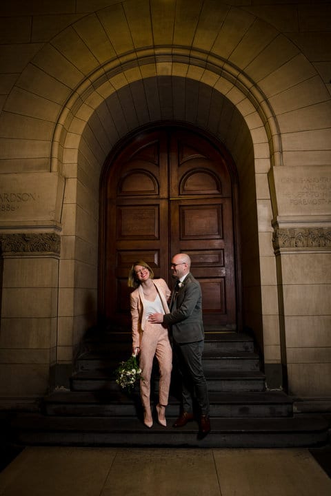 A couple share a laugh as they stand in front of a pair of large oak doors in a stone arch at the Allegheny County Courthouse.