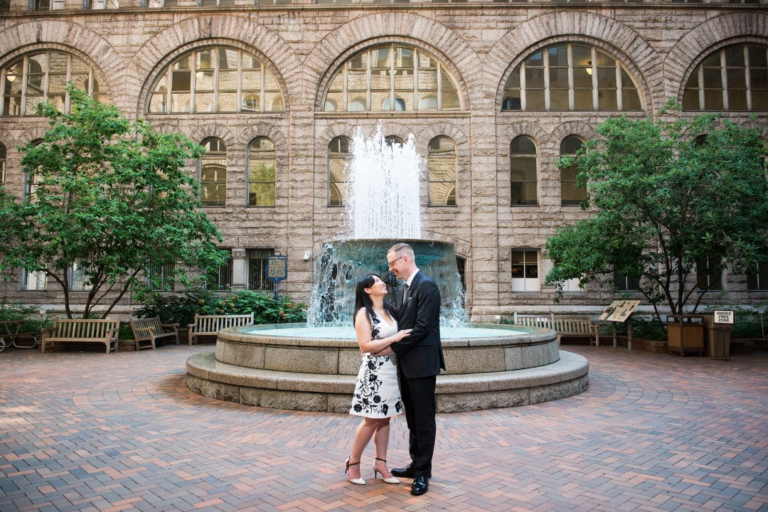 A bride and groom embrace in the courtyard of the Allegheny County Courthouse with the fountain behind them.