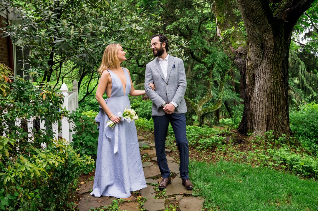 A bride and groom stand in on a wooded path. She's wearing a light blue dress and he's wearing a gray sport coat.