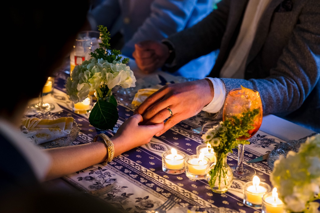 Closeup photo of a bride and groom holding hands across their banquet table lit by candles during their outdoor intimate wedding.