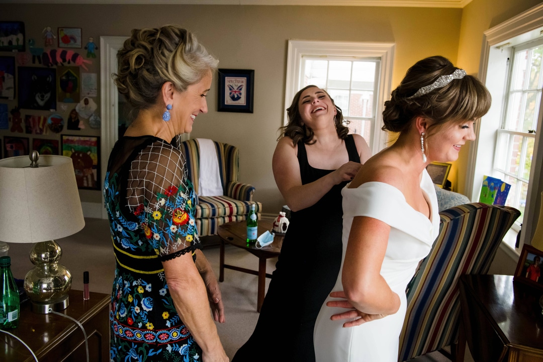 Two women laugh as they help a bride get dressed.