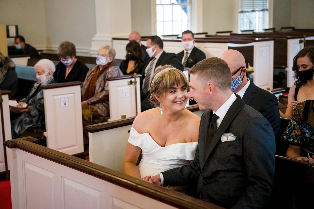 A bride and groom smile at each other while sitting in the first pew at a church.