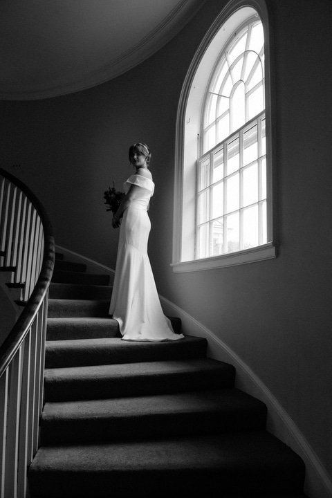 A bride stands next to a large window on a stairway.