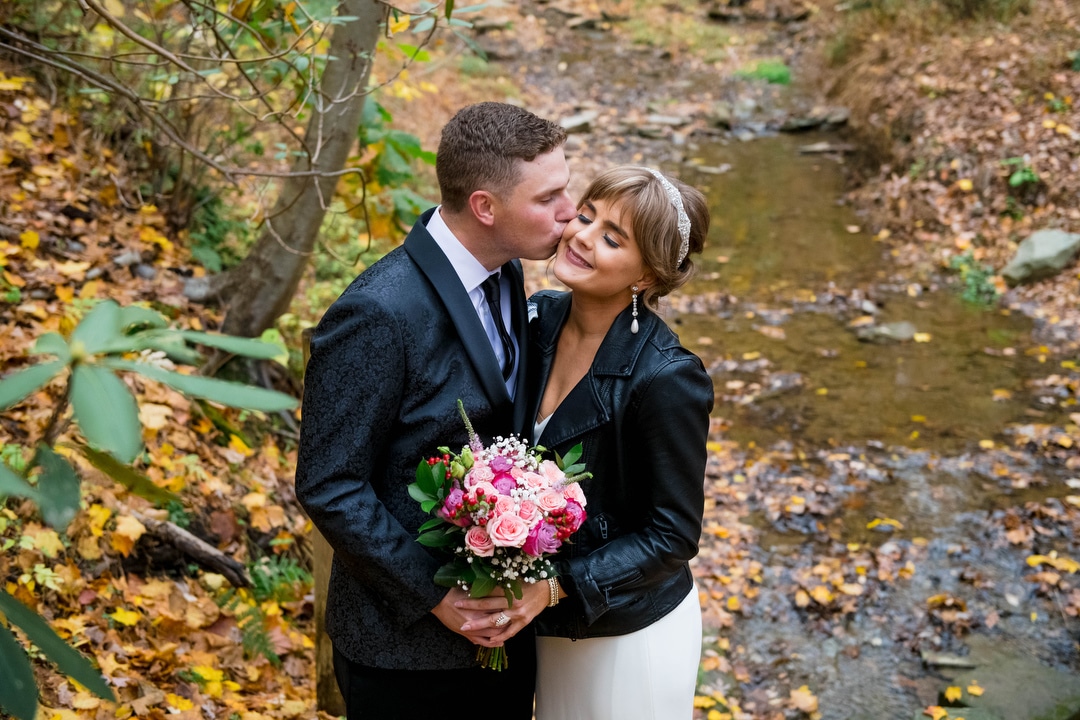 A groom kisses his bride's cheek as they stand by a stream in a forest.