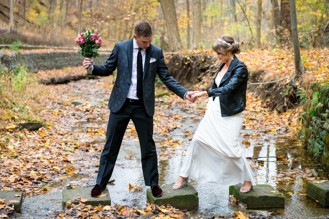 A groom helps his bride cross a stream on stepping stones.