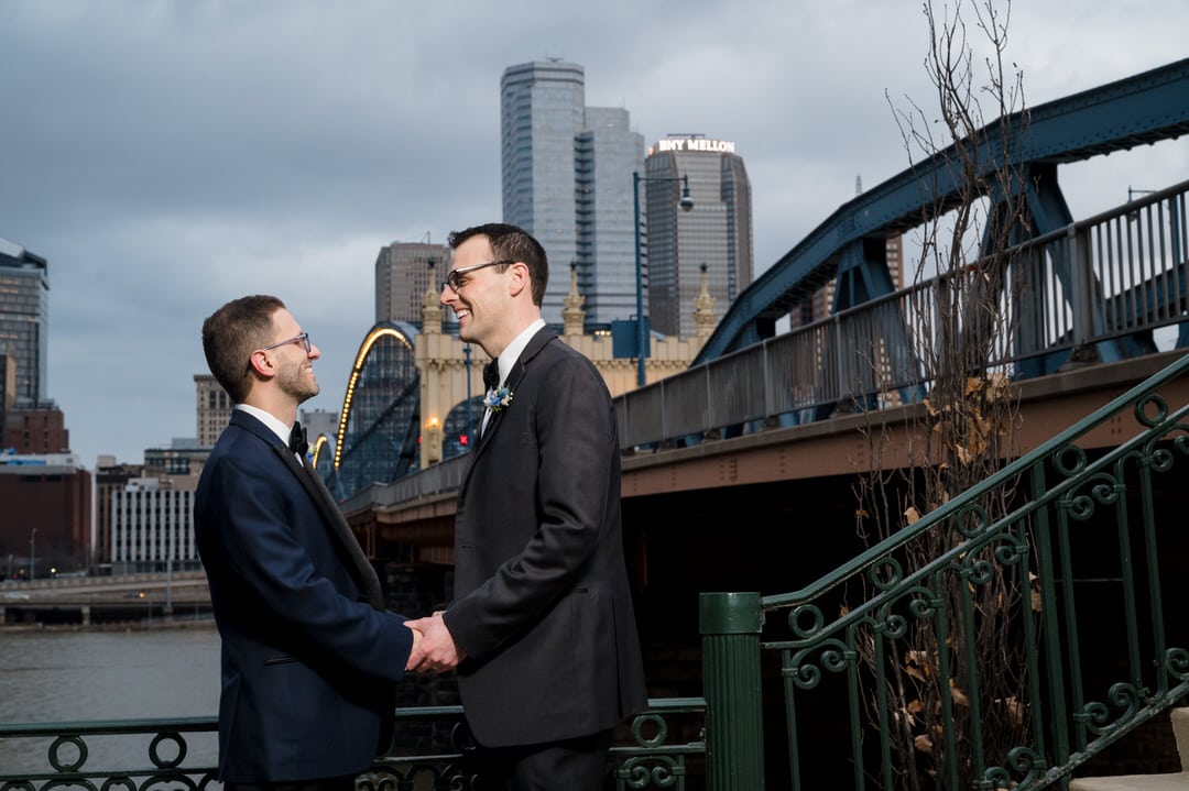 Two grooms hold hands in front of the Smithfield Street Bridge in Pittsburgh